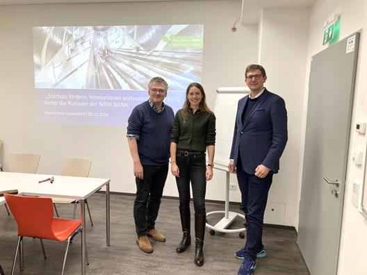Sebastian Hanny-Busch , Antonia Nörthemann und Prof. Dr. Olexiy Khabyuk stehen am Kopf eines Seminarraums. Hinter Ihnen wird das Startbild der Präsentation zum Gastvortrag an die Wand gebeamt.